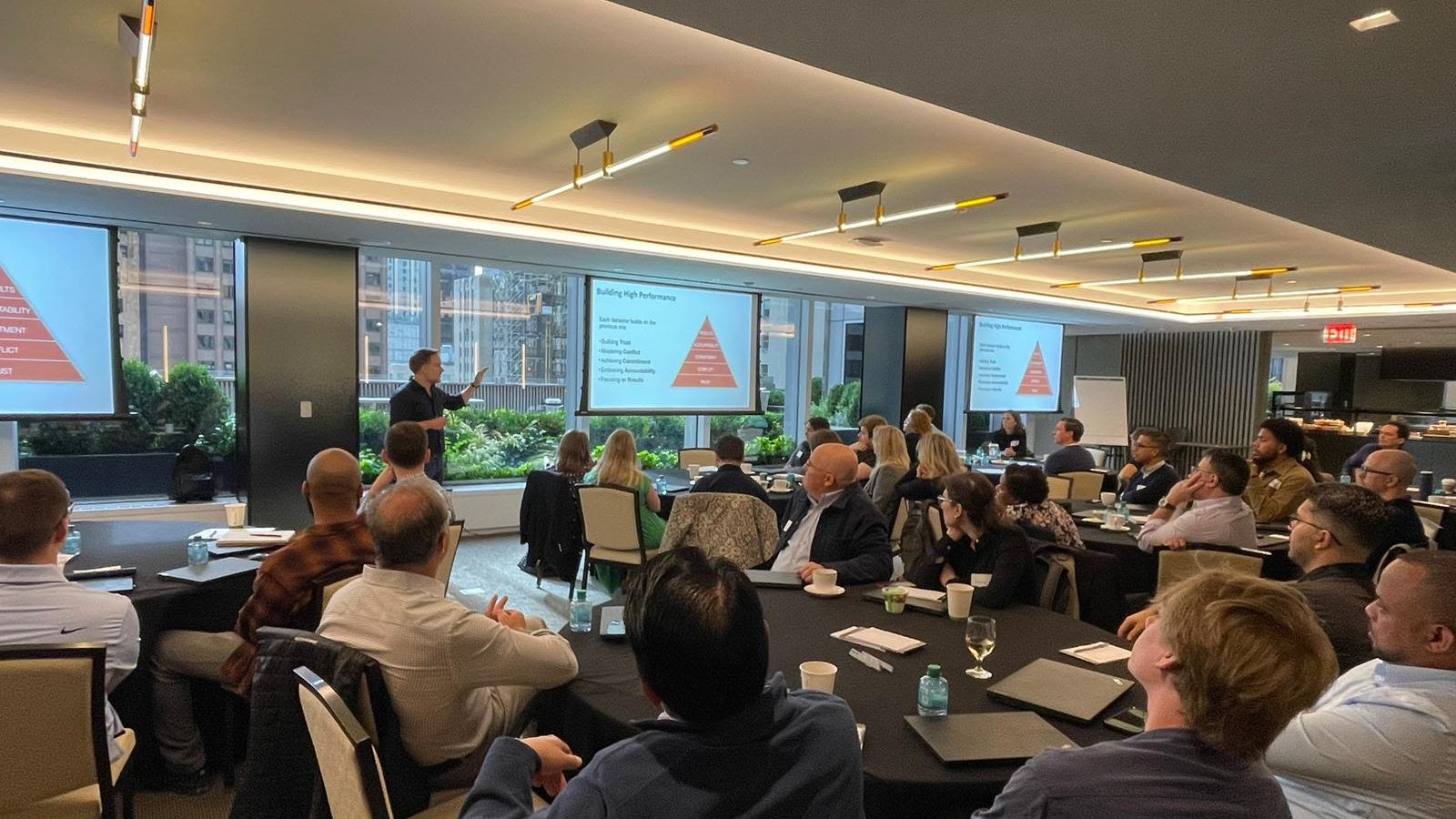 David O'Grady standing to present to a seated leadership team around boardroom tables, two wall screens showing the red Five Behaviors pyramid